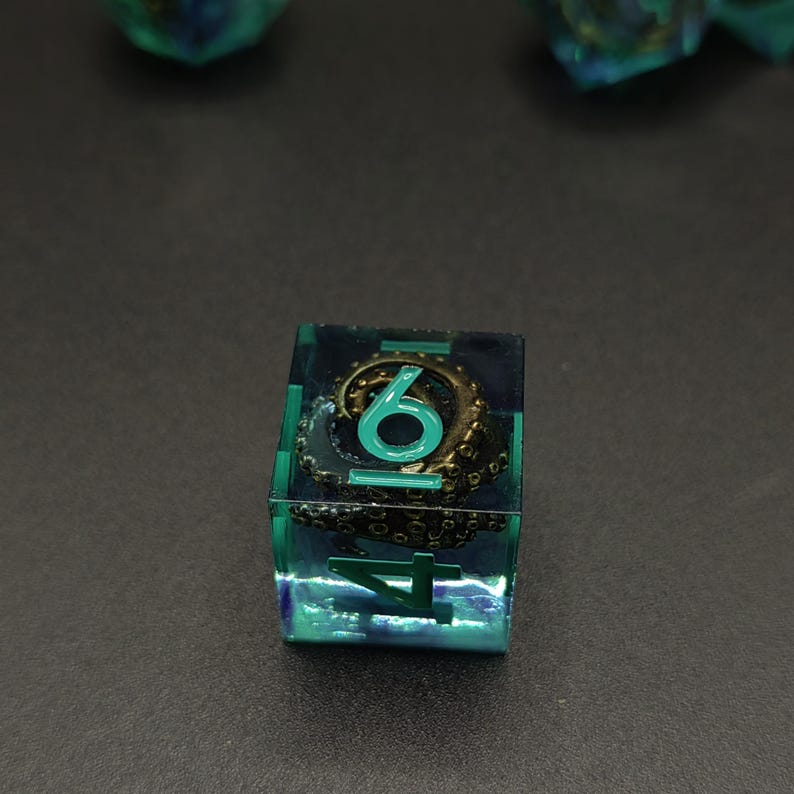 May include: A translucent teal and blue square die featuring an embedded metallic bronze octopus tentacle. The die displays the numbers 4 and 6 in a bright teal font, with the tentacle curling around the numeral 6 inside the resin.