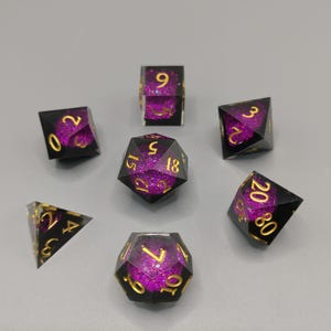 Peut inclure: Un ensemble de sept dés polyédriques pour jeux de rôle. Chaque dé est noir avec des chiffres dorés et un intérieur scintillant violet. L'ensemble comprend un d4, d6, d8, d10, d12, d20 et un d100.