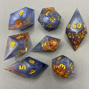 May include: A set of seven polyhedral dice in various shapes. The dice are a translucent blue with gold-colored numbers and gold leaf inclusions. The dice are designed for tabletop role-playing games.