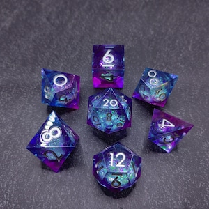 May include: Set of seven polyhedral dice in shades of purple, blue, and green. The dice have a glittery, galaxy-like appearance with white numbering. The dice are arranged on a dark gray surface.