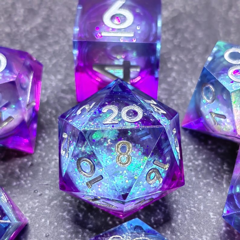 May include: Close-up of several polyhedral dice with a shimmering, iridescent effect. The dice are primarily blue and purple, with visible numbers in a silver font. The dice are designed for tabletop role-playing games.