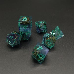 May include: A set of polyhedral dice made of translucent teal and purple resin. Each die contains a miniature bronze-colored octopus tentacle sculpture inside. The numbers on the dice are painted in a bright teal color for visibility.