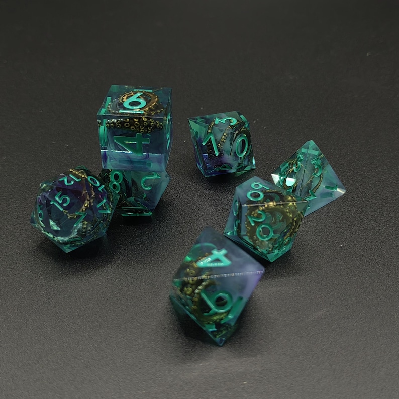 May include: A set of polyhedral dice made of translucent teal and purple resin. Each die contains a miniature bronze-colored octopus tentacle sculpture inside. The numbers on the dice are painted in a bright teal color for visibility.