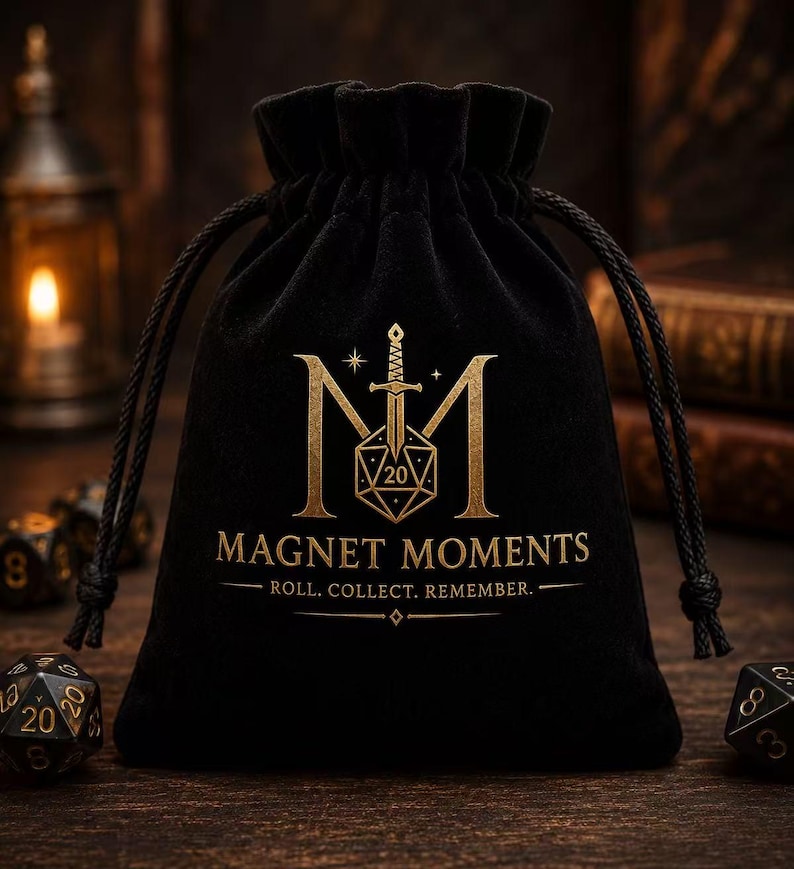 May include: A black velvet drawstring pouch featuring a gold-colored logo with a sword, a twenty-sided die, and the text MAGNET MOMENTS ROLL. COLLECT. REMEMBER. The bag is cinched at the top with black cords and sits on a dark wooden surface.