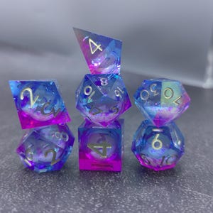 May include: A set of seven polyhedral dice in shades of blue and purple with gold numbering. The dice are transparent with glitter and a cloudy interior. The dice are arranged in a stack.