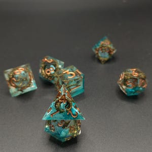 Puede incluir: Un conjunto de siete dados poli&eacute;dricos con tem&aacute;tica de fantas&iacute;a. Los dados son de color turquesa y transparentes con detalles y n&uacute;meros de color cobre. Los dados est&aacute;n dise&ntilde;ados para juegos de rol de mesa.