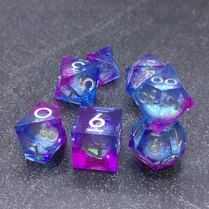 May include: A set of seven polyhedral dice in shades of blue and purple. The dice are transparent with glitter and gold numbering. The dice are for tabletop games.