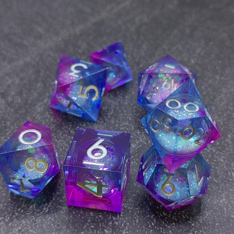 May include: A set of seven polyhedral dice in shades of blue and purple with gold numbering. The dice are transparent with glitter suspended inside, creating a shimmering effect. The dice are designed for tabletop role-playing games.
