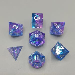 Puede incluir: Un conjunto de siete dados poliédricos de varias formas, incluyendo un cubo, d4, d6, d8, d10, d12 y d20. Los dados son una mezcla de azul y morado con purpurina iridiscente y numeración blanca. Los dados son para juegos de mesa.