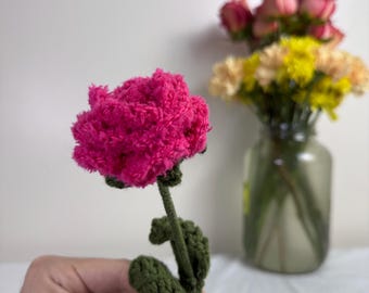 May include: A handmade, bright pink crochet flower with a green stem and leaves. The flower is held in a hand, with a blurred background of other flowers in a glass jar. The crochet flower is a unique, handcrafted item.