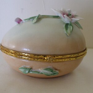 May include: A porcelain egg-shaped trinket box with a hinged lid. The box is decorated with a white flower with pink accents and green leaves. The lid has a gold trim.