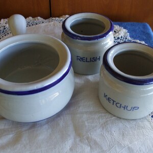 May include: Three white ceramic condiment jars with blue trim. The jars are labeled "Relish", "Ketchup", and one is empty. The jars are on a white tablecloth with a lace overlay.