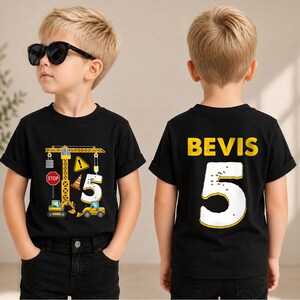 May include: A black short-sleeved t-shirt featuring a construction-themed graphic on the front with a yellow crane, excavators, a stop sign, and the number 5. The back displays the name BEVIS in yellow letters above a large white number 5.