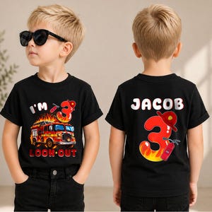 May include: A black t-shirt featuring a fire truck graphic with flames, the text I'M 3 LOOK OUT, and a large number 3 with a fire helmet. The back displays the name JACOB above a large number 3, a fire helmet, and a fire extinguisher graphic.