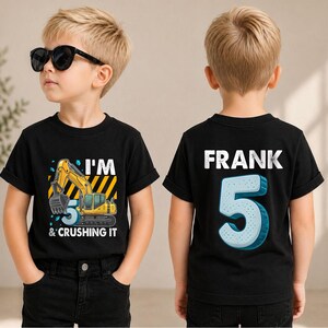 May include: A black short-sleeved t-shirt featuring a yellow excavator graphic with the text I'M 5 & CRUSHING IT on the front and the name FRANK with a large number 5 printed in white and light blue on the back of the garment.