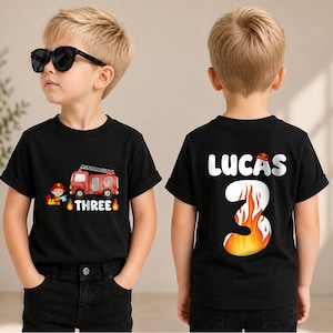 May include: A black short-sleeved t-shirt featuring a graphic of a red fire truck, a firefighter, and the word THREE on the front. The back displays the name LUCAS above a large number 3 filled with a flame pattern and a small fire helmet.