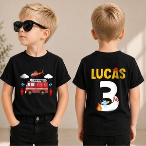 May include: A black short-sleeved t-shirt featuring a graphic of a red fire truck and helicopter on the front. The back displays the name LUCAS in yellow letters above a large white number 3, accented with a small fire helmet and water hose.