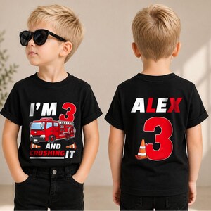 May include: A black t-shirt featuring a red fire truck graphic with the text I'M 3 AND CRUSHING IT on the front, and the name ALEX with a large red number 3 and a traffic cone graphic on the back, paired with black denim jeans.