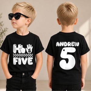 May include: A black short-sleeved t-shirt featuring a white graphic on the front that reads "Hi Five" with a hand icon and a checkered pattern. The back of the shirt displays the name "ANDREW" above a large white number "5" in a bold font.
