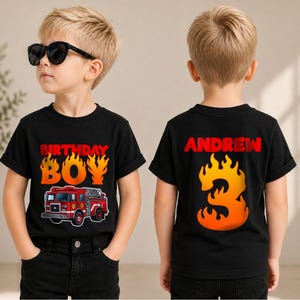May include: A black short-sleeved t-shirt featuring a red fire truck graphic and the text "BIRTHDAY BOY" in orange and yellow flames on the front, and the name "ANDREW" above a large orange and yellow flame-shaped number "3" on the back.