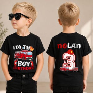 May include: A black t-shirt featuring a red fire truck graphic with the text I'M THE BIRTHDAY BOY on the front and the name NOLAN with a large number 3 and a fire truck graphic on the back, shown from front and back views.