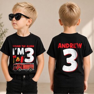 May include: A black short-sleeve t-shirt featuring a firefighter theme. The front displays the text "SOUND THE ALARM I'M 3" with a fire truck, axe, and fire graphic. The back features the name "ANDREW" above a large number 3 in white.