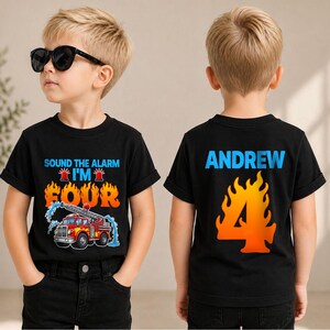 May include: A black t-shirt featuring a graphic of a red fire truck with blue water, orange flames, and the text "SOUND THE ALARM I'M FOUR" on the front, and the name "ANDREW" above a large orange flame-patterned number "4" on the back.