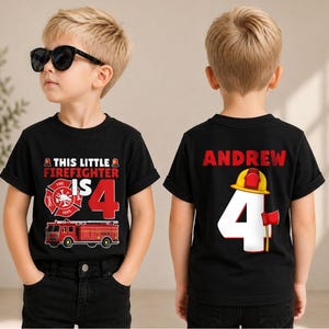 May include: A black t-shirt featuring a firefighter theme. The front displays a red fire truck, a fire department emblem, and the text "THIS LITTLE FIREFIGHTER IS 4". The back shows the name "ANDREW" above a large white number 4 and a yellow helmet.