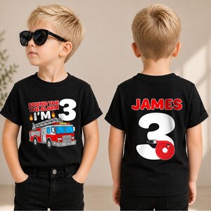 May include: A black short-sleeved t-shirt featuring a fire truck graphic with the text 'SOUND THE ALARM I'M 3' on the front and the name 'JAMES' above a large number '3' with a red fire hose graphic on the back.
