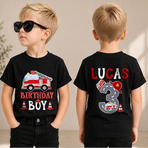 May include: A black t-shirt featuring a red fire truck graphic, the text "BIRTHDAY BOY" in red and white, and orange traffic cones. The back displays the name "LUCAS" in red, a large gray number "3" with a road pattern, a fire truck, and a helmet.