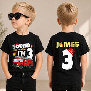 May include: A black short-sleeved t-shirt featuring a graphic of a red fire truck and the text "SOUND THE ALARM I'M 3" on the front, and the name "JAMES" with a large white number "3" and a red firefighter helmet graphic on the back.