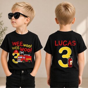 May include: A black t-shirt featuring a graphic print of a red fire truck, a yellow number 3, a red fire helmet, and the text WEE WOO! WEE WOO! I'M 3 on the front, with the name LUCAS, a yellow number 3, and a red fire extinguisher on the back.