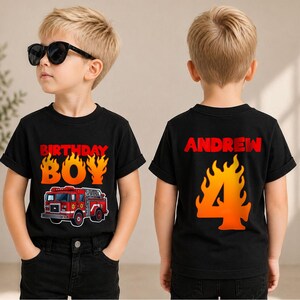 May include: A black short-sleeved t-shirt featuring a red fire truck graphic and the text "BIRTHDAY BOY" in orange flames on the front, and the name "ANDREW" above a large orange flame-patterned number "4" on the back of the garment.