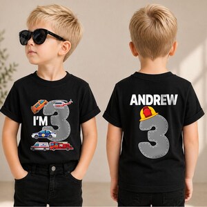 May include: A black short-sleeved t-shirt featuring a graphic of a gray number 3 with emergency vehicles and the text I'M 3 on the front, and the name ANDREW above a gray number 3 with a yellow and red firefighter helmet on the back.