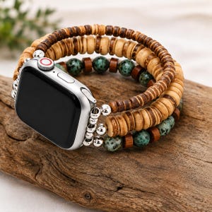 Może przedstawiać: Srebrny smartwatch z czarnym ekranem przymocowany do wielowarstwowej bransoletki. Bransoletka składa się z rzędów jasnych i ciemnych drewnianych koralików w kształcie dysków, przeplatanych małymi srebrnymi metalowymi koralikami oraz okrągłymi, nakrapianymi turkusowymi kamieniami.
