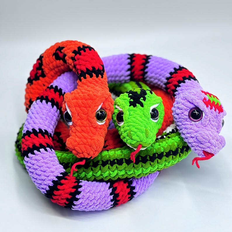 Peut inclure: Trois serpents crochet&eacute;s aux couleurs vives s'enroulent ensemble. L'un est orange et rouge, un autre vert, et le troisi&egrave;me violet et rouge. Chaque serpent a de grands yeux en bouton et une langue rouge, cr&eacute;ant un affichage ludique et color&eacute;.