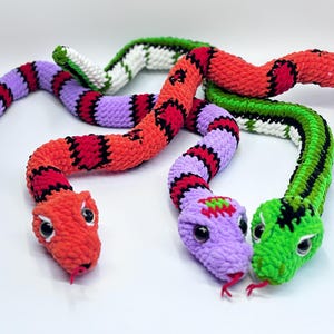 Peut inclure: Trois serpents en crochet color&eacute;s, rouge, violet et vert, avec des accents noirs et blancs. Chaque serpent a une t&ecirc;te d&eacute;taill&eacute;e avec de grands yeux et une petite langue rouge. Les serpents sont dispos&eacute;s sur une surface blanche.