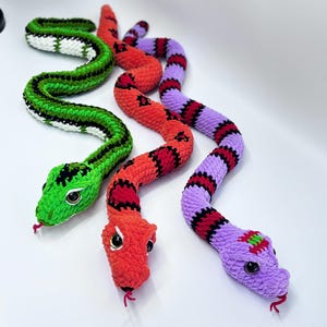Peut inclure: Trois jouets serpents crochet&eacute;s aux couleurs vives. L'un est vert avec des rayures noires et blanches, un autre est rouge avec des marques noires, et le troisi&egrave;me est violet avec des rayures rouges et noires. Chacun a de grands yeux ronds et une langue rouge.
