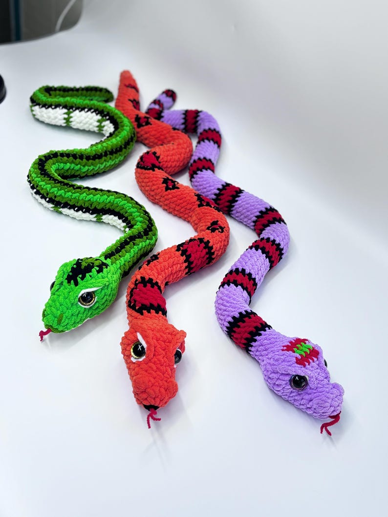 Peut inclure: Trois jouets serpents crochet&eacute;s aux couleurs vives. L'un est vert avec des rayures noires et blanches, un autre est rouge avec des marques noires, et le troisi&egrave;me est violet avec des rayures rouges et noires. Chacun a de grands yeux ronds et une langue rouge.