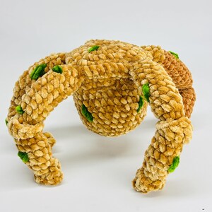 May include: A plush, handmade toy in a unique shape, featuring a central sphere with four curved appendages. The toy is primarily tan with small green accents, and has a soft, textured appearance. The toy is likely designed for children.