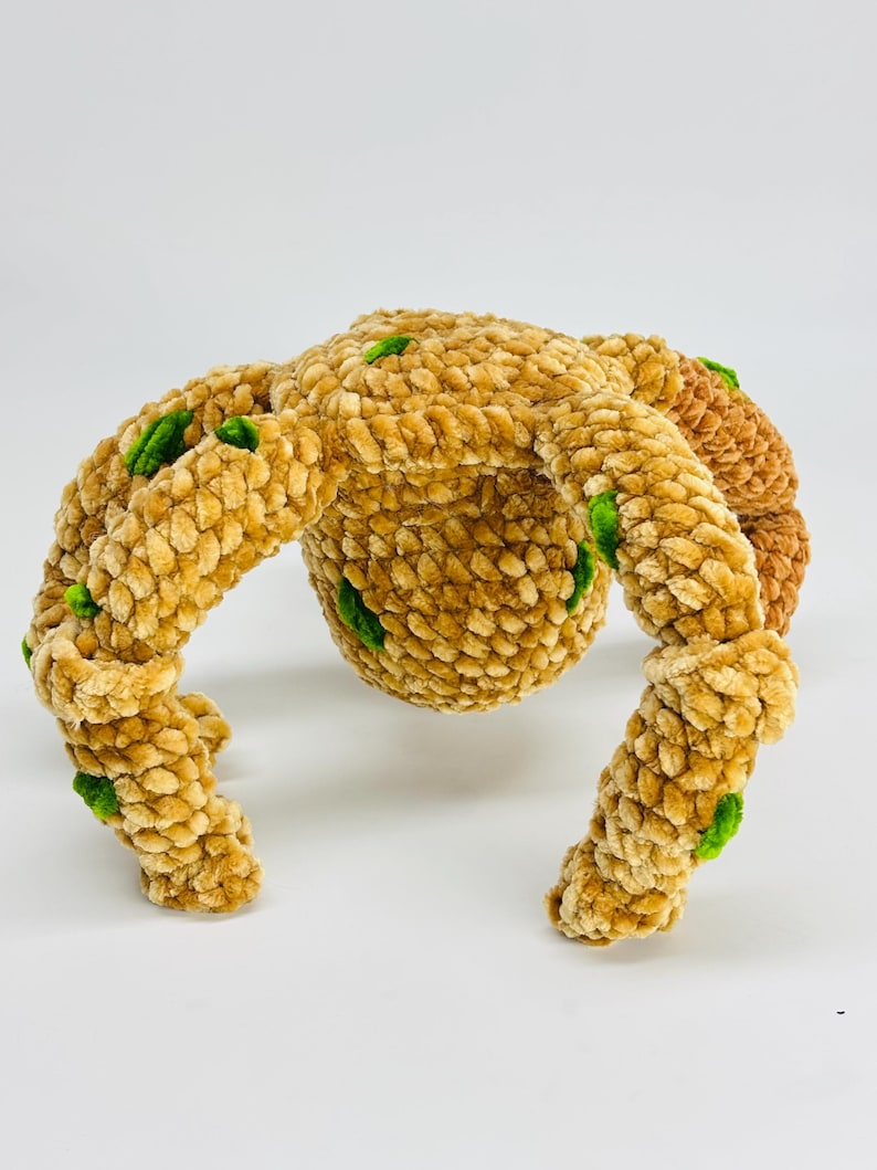 May include: A plush, handmade toy in a unique shape, featuring a central sphere with four curved appendages. The toy is primarily tan with small green accents, and has a soft, textured appearance. The toy is likely designed for children.