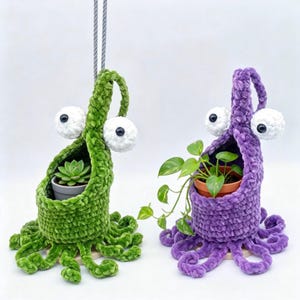 Può includere: Due originali portavasi all'uncinetto fatti a mano, verdi e viola. Ognuno presenta grandi occhi bianchi e sporgenti e appendici simili a tentacoli. Uno contiene una succulenta, l'altro una pianta verde e frondosa, sospesi da una corda grigia.
