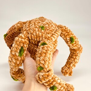 May include: A handmade, plush, brown monster toy with four limbs and a round body. The soft, textured toy has green accents and is held in a person's hand. The toy is made of a fuzzy material.