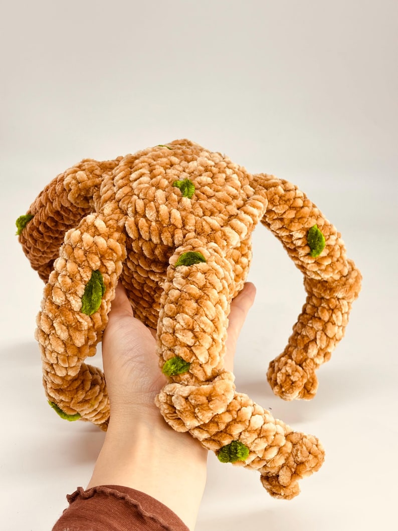 May include: A handmade, plush, brown monster toy with four limbs and a round body. The soft, textured toy has green accents and is held in a person's hand. The toy is made of a fuzzy material.