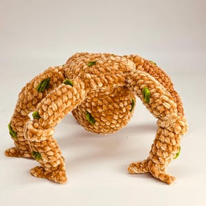 May include: A plush, brown, spider-like creature with eight segmented legs and a rounded body. The soft, textured toy has green accents and is set against a white background. The handmade toy is a unique, whimsical item.