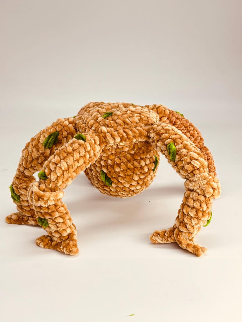 May include: A plush, brown, spider-like creature with eight segmented legs and a rounded body. The soft, textured toy has green accents and is set against a white background. The handmade toy is a unique, whimsical item.
