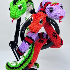Peut inclure: Sculpture de serpent crochet&eacute; avec trois t&ecirc;tes aux couleurs vives : vert, rouge et violet. Les serpents ont des yeux noirs et blancs et des langues rouges. La sculpture est mont&eacute;e sur un support noir.