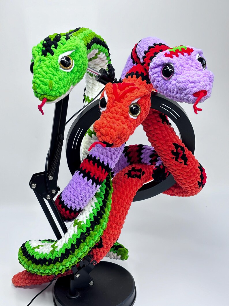Peut inclure: Sculpture de serpent crochet&eacute; avec trois t&ecirc;tes aux couleurs vives : vert, rouge et violet. Les serpents ont des yeux noirs et blancs et des langues rouges. La sculpture est mont&eacute;e sur un support noir.