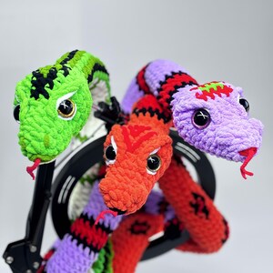 Peut inclure: Un serpent en crochet fait &agrave; la main, multicolore, avec trois t&ecirc;tes. Les t&ecirc;tes sont vertes, orange et violettes, chacune avec de grands yeux noirs et des langues rouges. Le serpent est mont&eacute; sur un support d'&eacute;clairage annulaire noir.