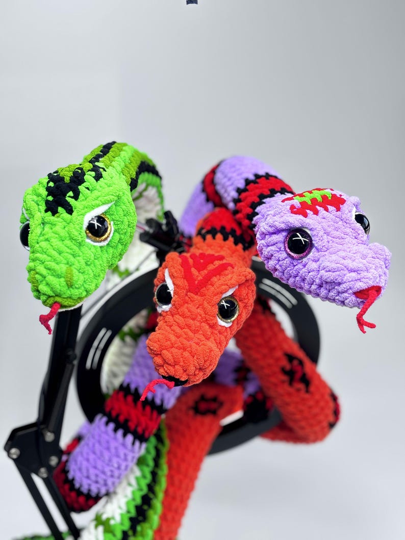 Peut inclure: Un serpent en crochet fait &agrave; la main, multicolore, avec trois t&ecirc;tes. Les t&ecirc;tes sont vertes, orange et violettes, chacune avec de grands yeux noirs et des langues rouges. Le serpent est mont&eacute; sur un support d'&eacute;clairage annulaire noir.